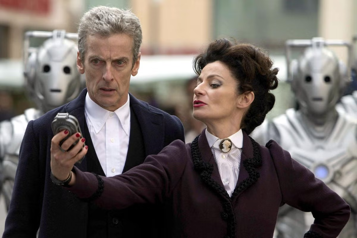 Missy leans into the Doctor's space, showcasing her device in a similar manner, arm outstretched, head tilted, red nailpolish visible in the flurishing hold. This is the Device with which she vapourises people, and she's showing the horrified doctor Her cyberman taking over the world. 
